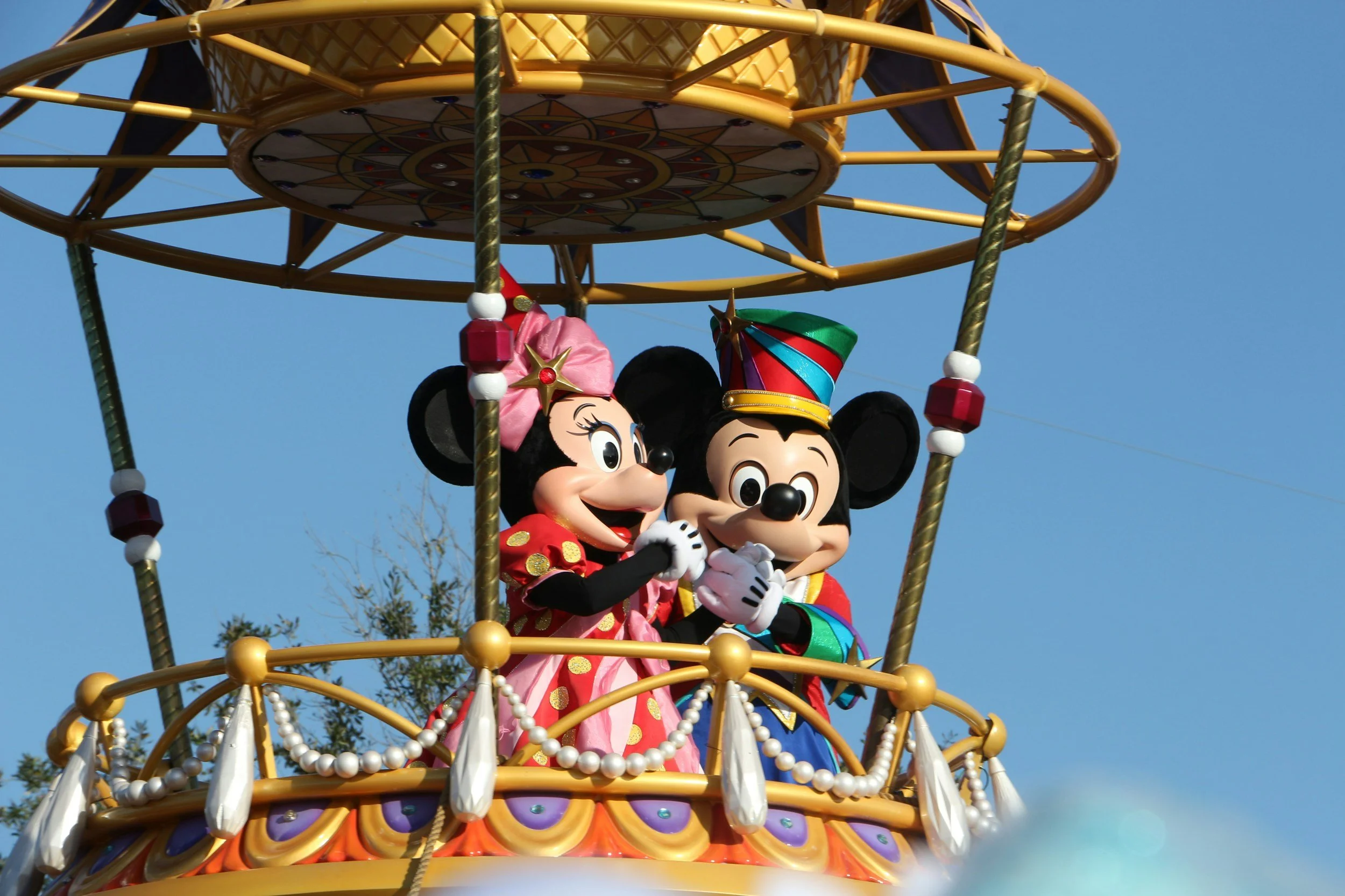 Minnie Mouse and Mickey Mouse in colorful costumes on a decorative float at a parade, with a clear blue sky in the background. Disney Dance Camp
