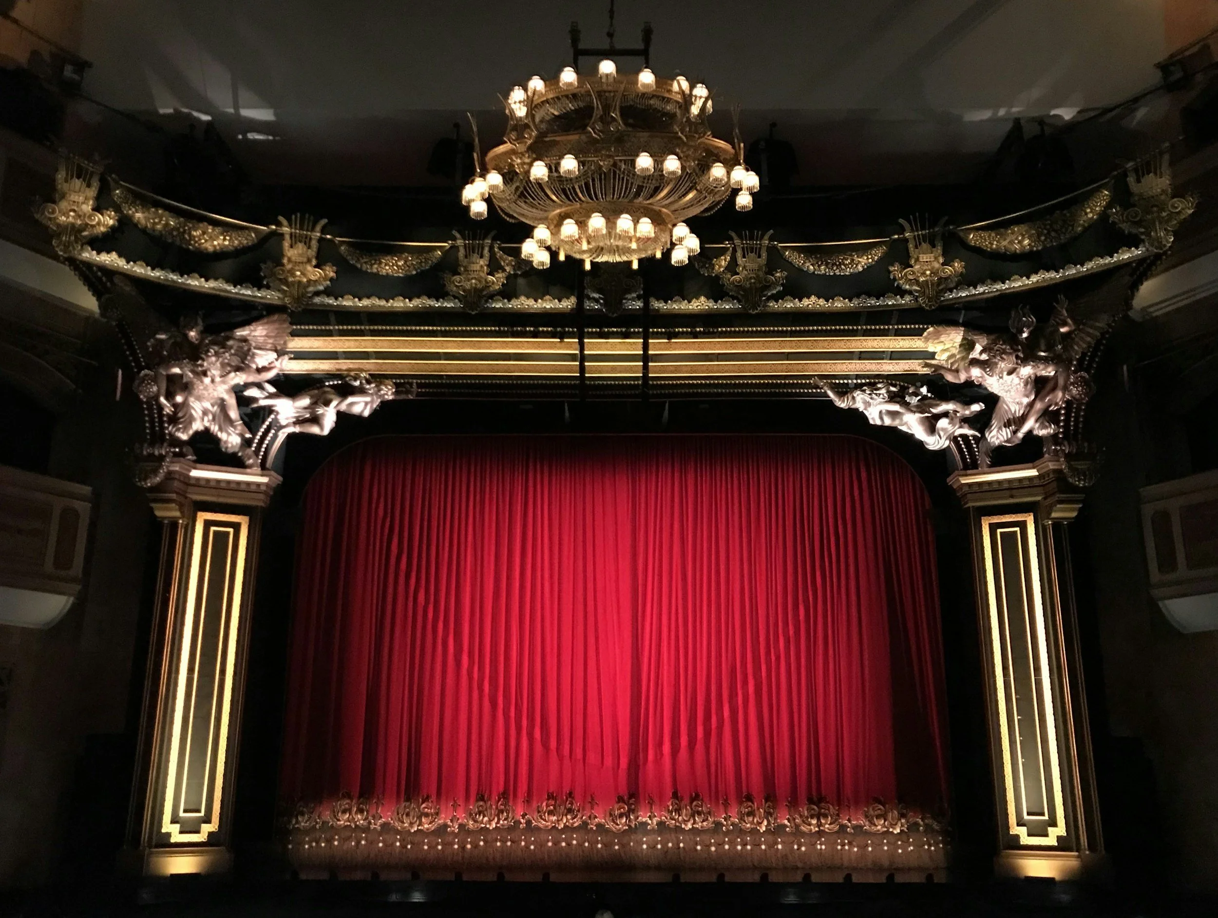 The image shows a theater stage with a closed red curtain, ornate gold and black pillars, and elaborate decorative sculptures, with a grand chandelier hanging from the ceiling. Musical Theater Dance Camp