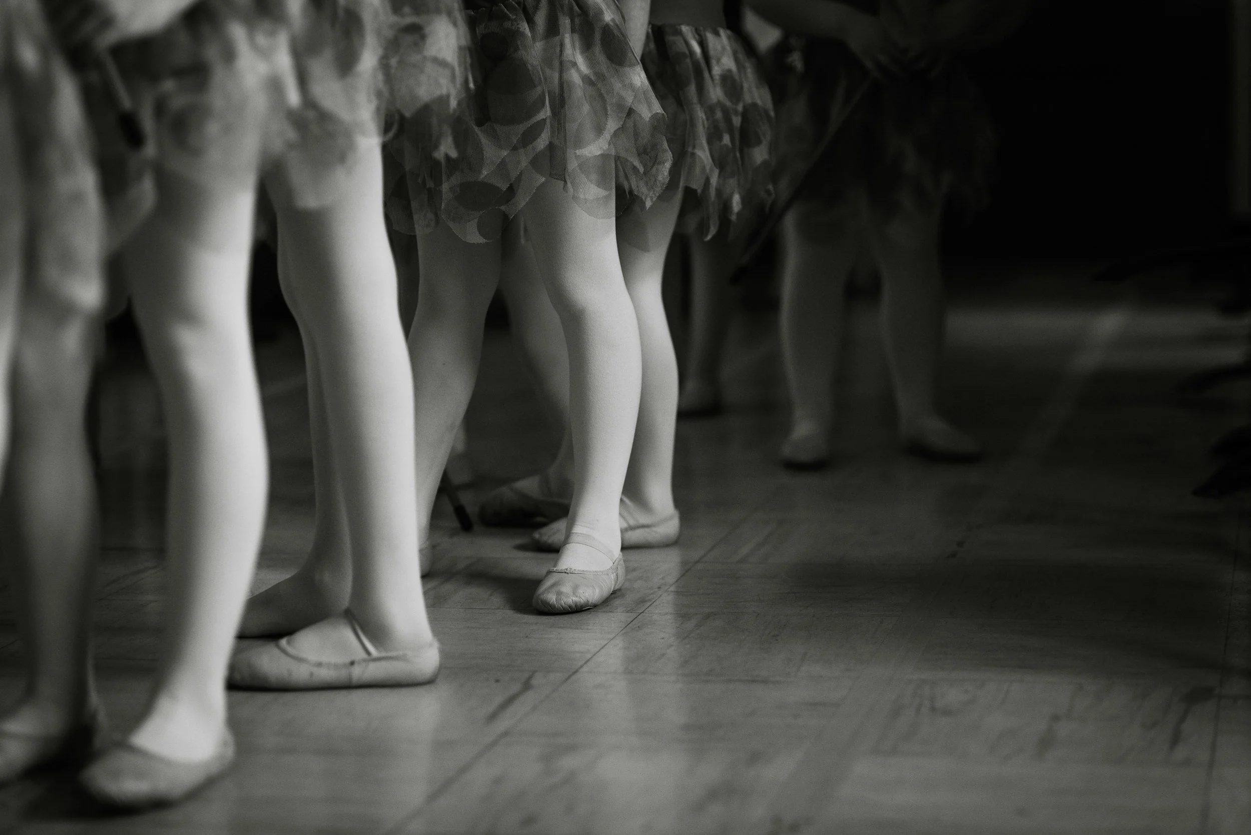 Several young girls wearing ballet slippers and tutus standing in a line on a wooden dance floor.