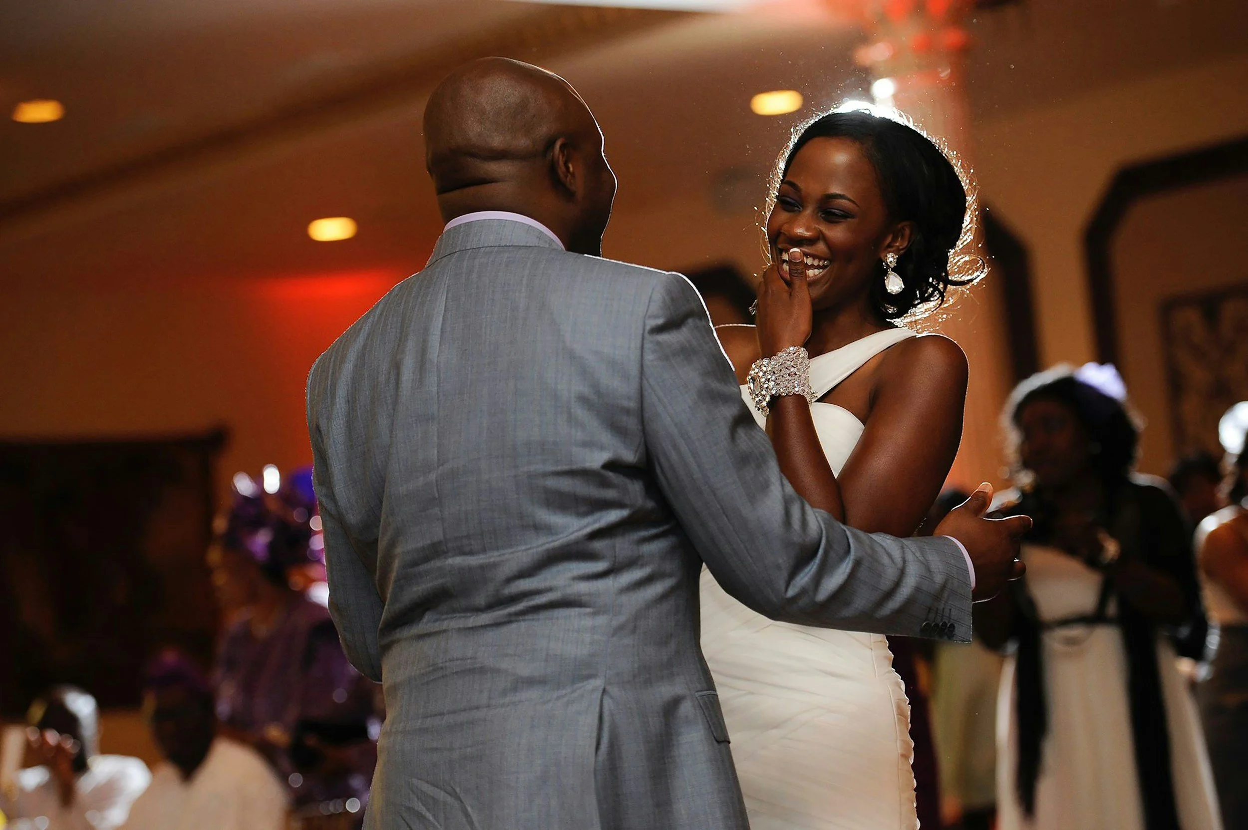 A bride and groom dancing at their wedding reception, smiling and enjoying a moment together. The bride is wearing a white dress with jewelry, and the groom is in a gray suit. The background shows other guests at the reception.