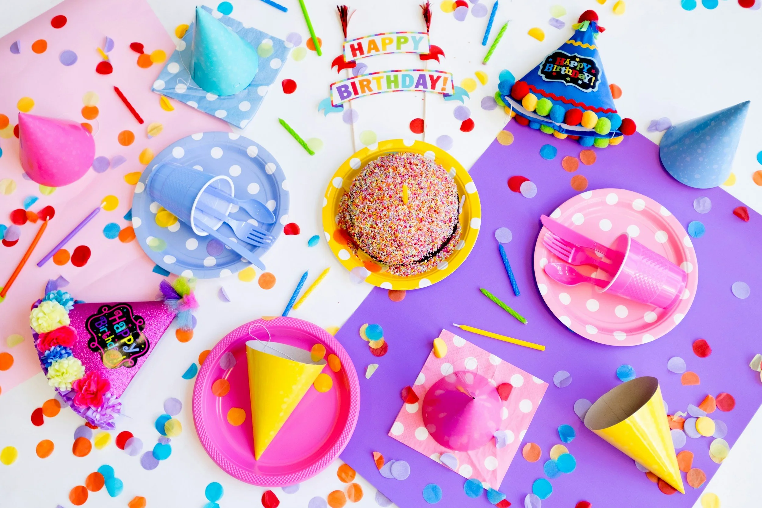 Colorful birthday party table with confetti, party hats, plates, cups, balloons, and a frosted birthday cake.