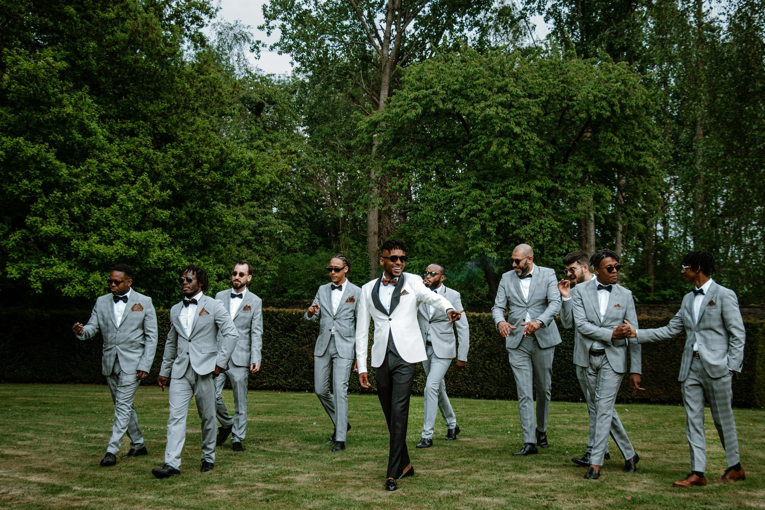 A group of men in matching gray suits with black bow ties and dress shoes, walking on a grassy area outdoors, with a man in a white tuxedo jacket leading and smiling, surrounded by men wearing sunglasses, with tall trees in the background.