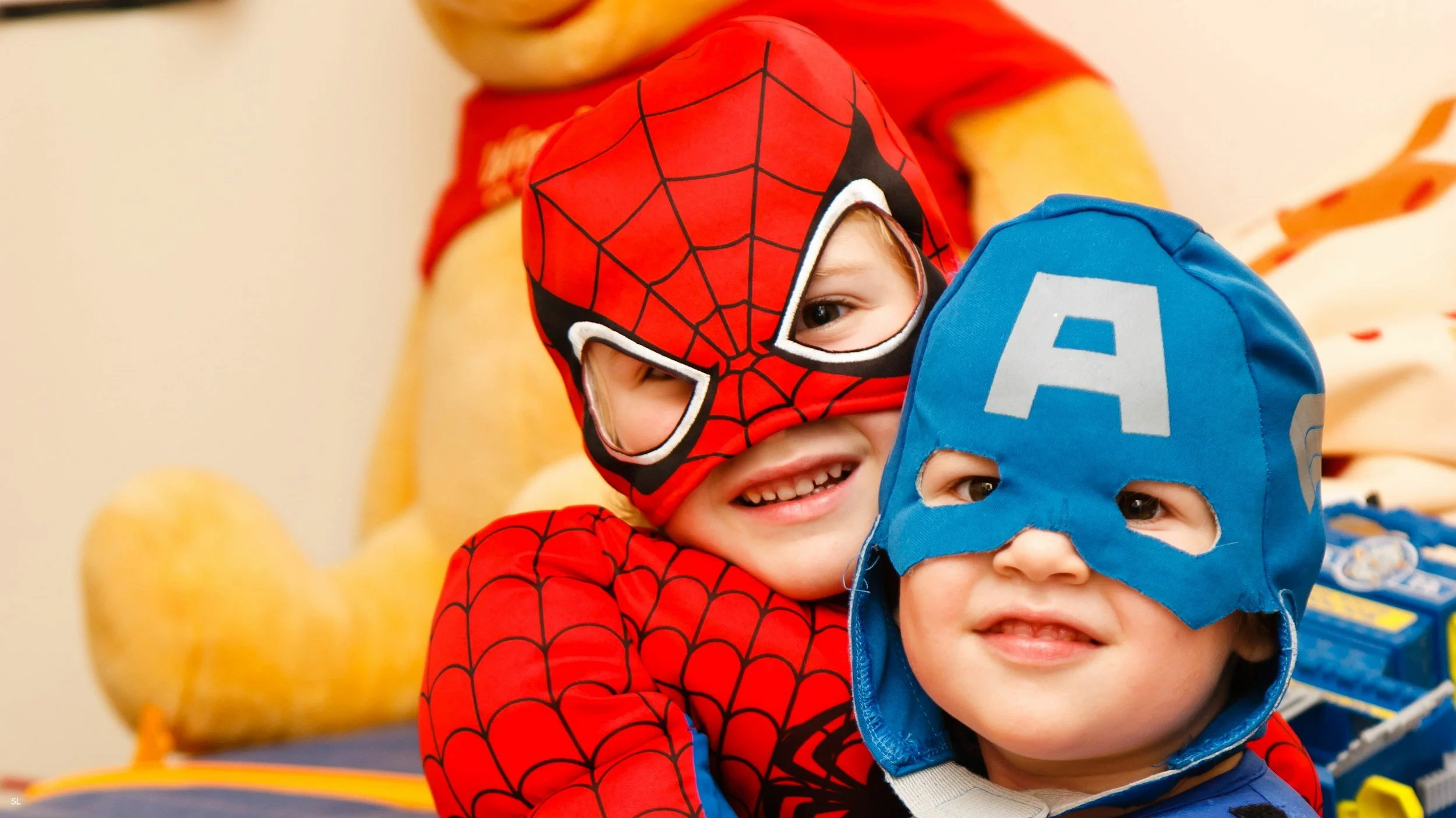 Two young children dressed as superheroes, Spider-Man and Captain America, smiling and posing together indoors.