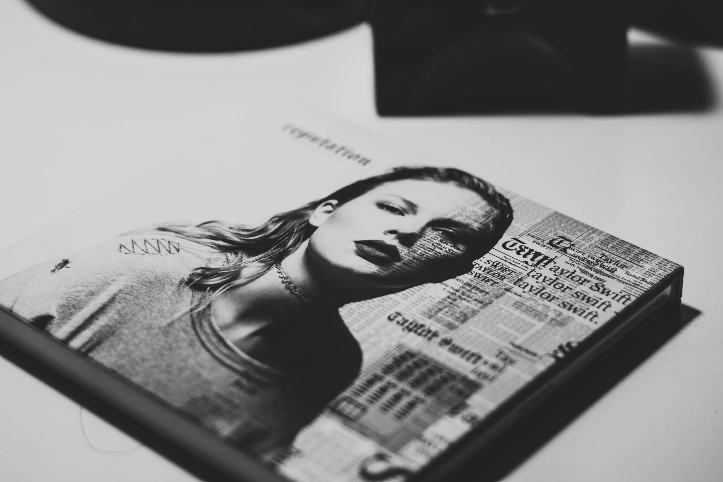 Black and white photo of a magazine featuring a portrait of a woman with long hair, dark lipstick, and jewelry, placed on a table with newspapers in the background. Dance Camp with Taylor Swift Music