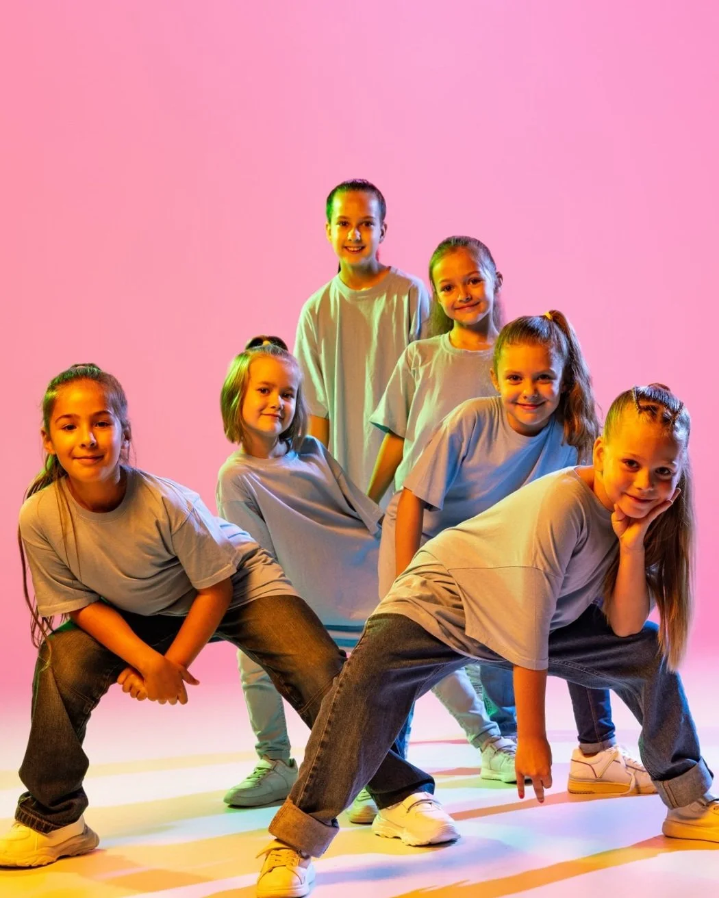 Six young girls wearing beige t-shirts and jeans posing against a pink background, with colorful lighting creating a vibrant scene. Hip Hop Dance