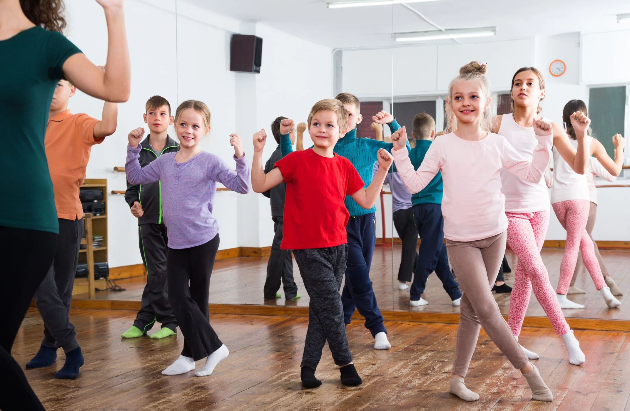 Children participating in a dance class in a studio, following an instructor, with a mirror reflecting their movements.