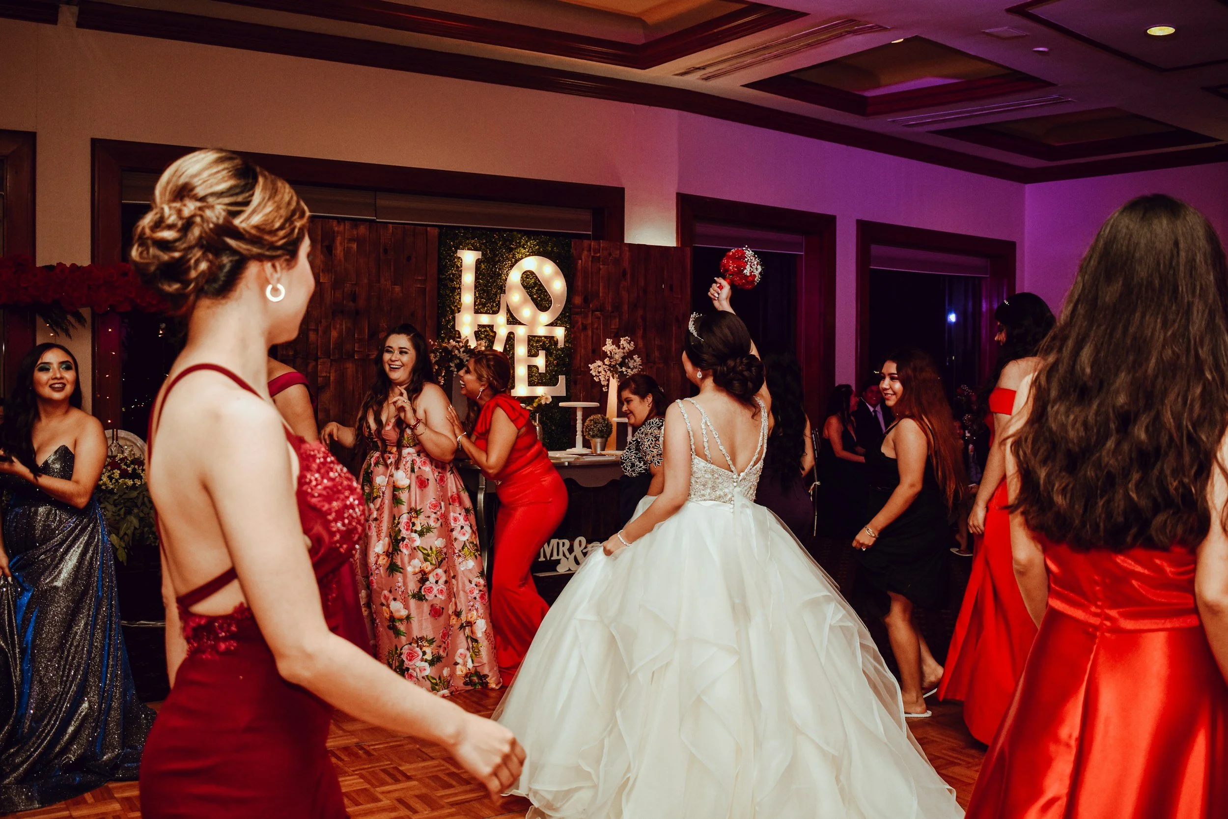 Group of women dancing at a wedding reception in a decorated ballroom, with a bride in a white wedding gown, a large illuminated 'LOVE' sign in the background, and guests in colorful dresses.