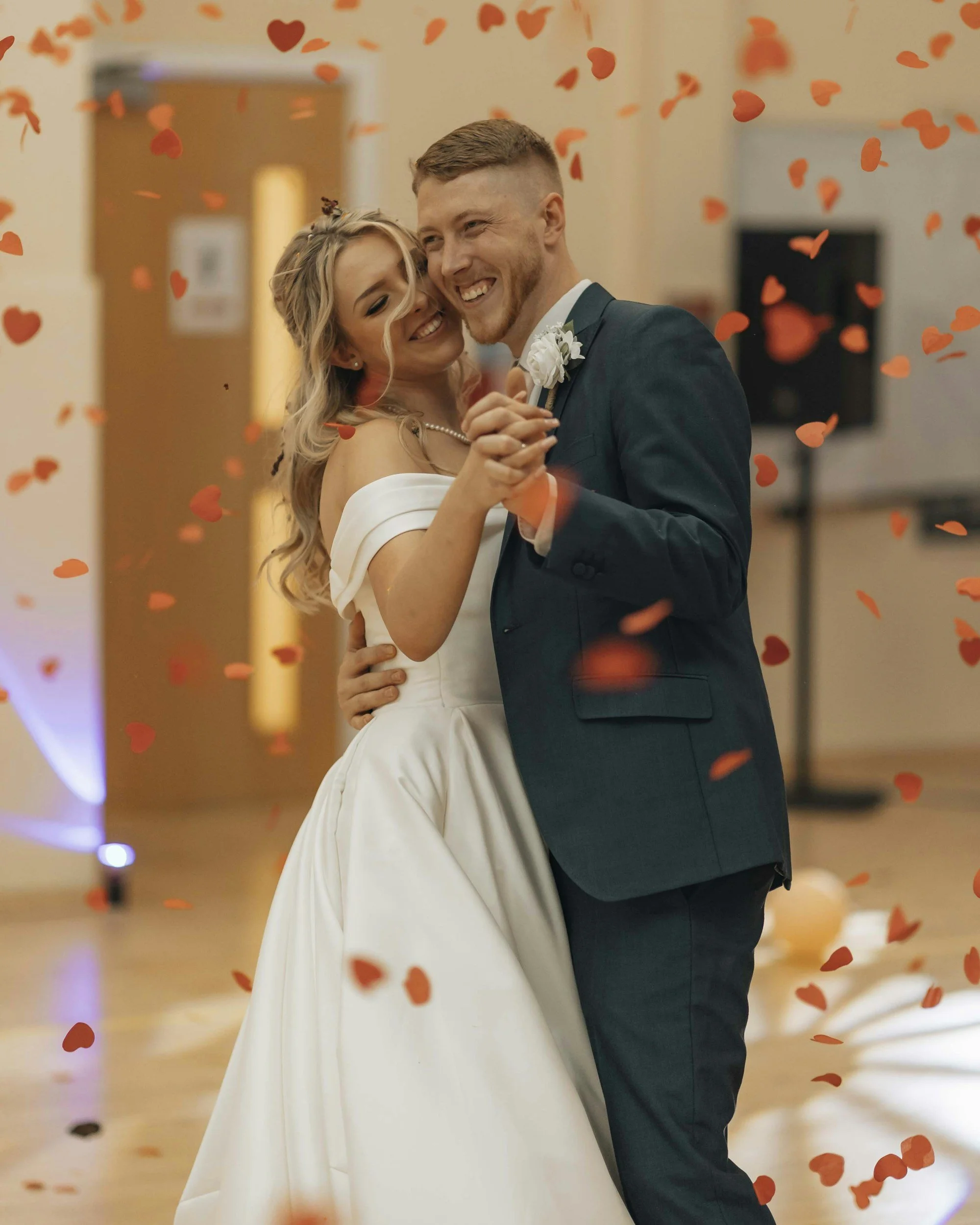 A bride and groom dancing and smiling during their wedding reception, surrounded by orange heart-shaped confetti.