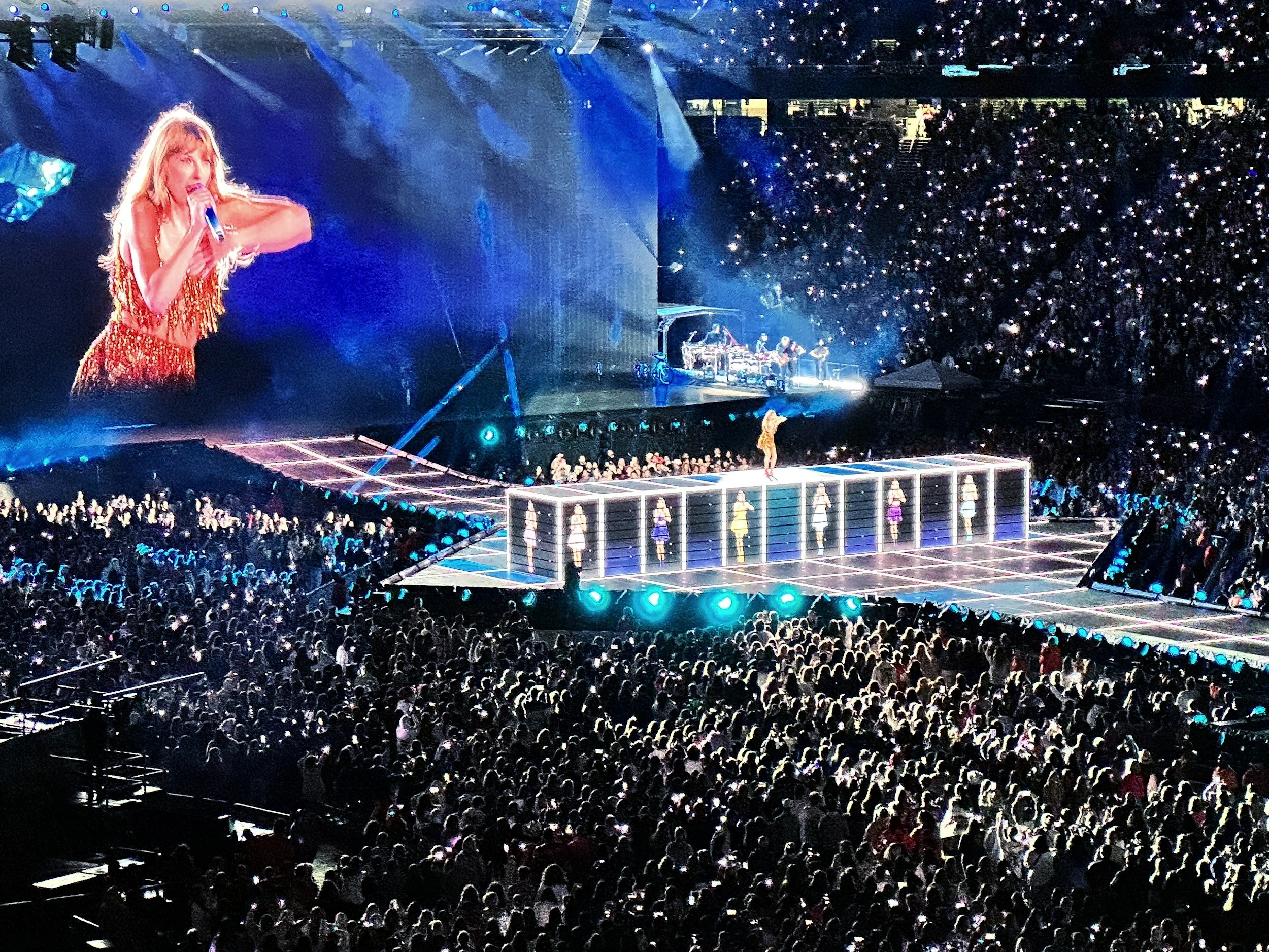 Concert stage with singer performing, large screen displaying close-up of singer, audience with many lights, and backup dancers on stage. Taylor Swift Camp
