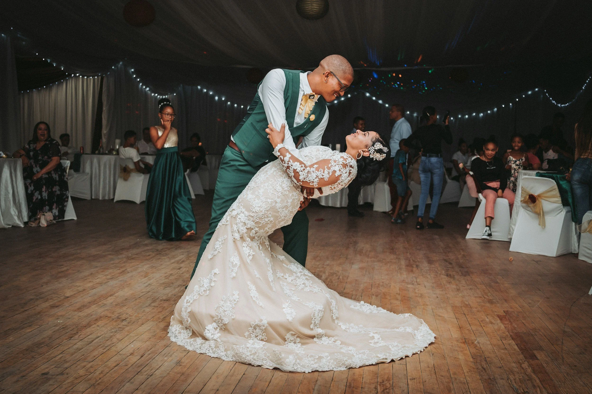 A bride and groom dancing at their wedding reception, with the groom dipping the bride backwards on a wooden dance floor. The bride is in a white lace wedding gown with long sleeves, and the groom is in a green vest and trousers with a light shirt and tie. Guests are seated and standing around them, watching and smiling.